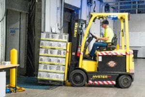 A worker drives a Hyster forklift moving Joe IPA boxes in a warehouse.